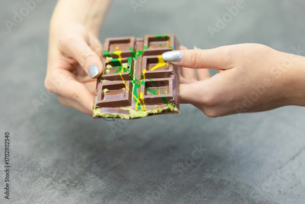 Obraz Female hands breaking off piece of Dubai chocolate on gray background. Green kadayif and pistachio spread with pistachio in trend dessert. Close-up, selective focus. High quality photo