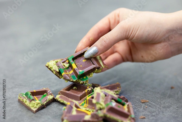 Obraz Woman's hand holds piece of Dubai chocolate on gray background. Green kadayif and pistachio spread with pistachio in trend dessert. Close-up, selective focus. High quality photo