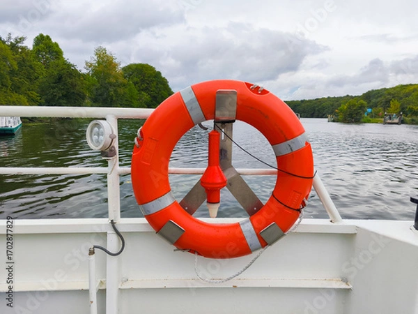 Obraz Berlin, Germany - September 05, 2025: Bright orange life buoy mounted on a boat railing, surrounded by tranquil water and lush green trees, emphasizing safety on the water