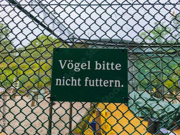 Obraz Green sign with white text instructing visitors not to feed birds, displayed on a wire fence in a nature reserve, surrounded by lush greenery and natural habitat