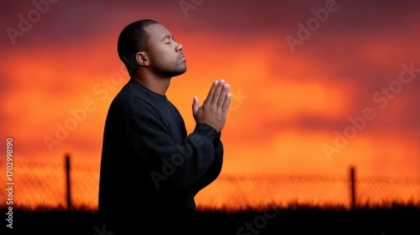 Fototapeta Thoughtful Man in Prayer Against a Vibrant Sunset Background with Warm Colors and Atmospheric Clouds