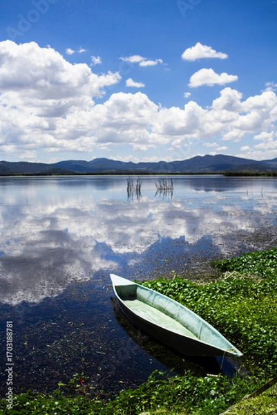 Fototapeta Small wooden fishing boat resting on the calm water of a lake, reflecting the beautiful cloudy sky and surrounding mountains