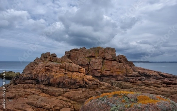 Fototapeta Massive reddish coastal cliffs covered with moss and lichen under a dramatic cloudy sky. Wild seashore landscape with textured rocks and ocean in the background. France