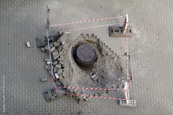 Fototapeta Urban Maintenance Site with Manhole Cover and Safety Barriers – Infrastructure Repair Scene. A circular manhole cover is surrounded by displaced paving bricks and cordoned off with red-white tape.