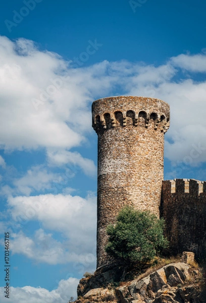 Fototapeta Ancient stone tower standing tall against a bright blue sky with white clouds, showcasing the historic architecture of Tossa de Mar in Costa Brava, Spain.