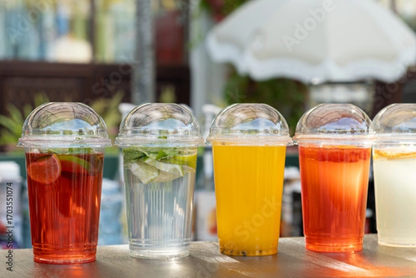 Fototapeta Row of assorted refreshing beverages in plastic cups with dome lids, displayed on a wooden surface at a summer street cafe or outdoor market