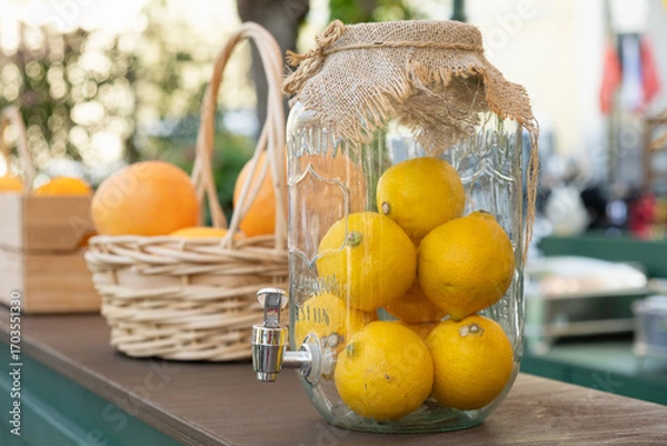Fototapeta Large glass beverage dispenser filled with fresh lemons, decorated with burlap on top, displayed at a countryside market or fair