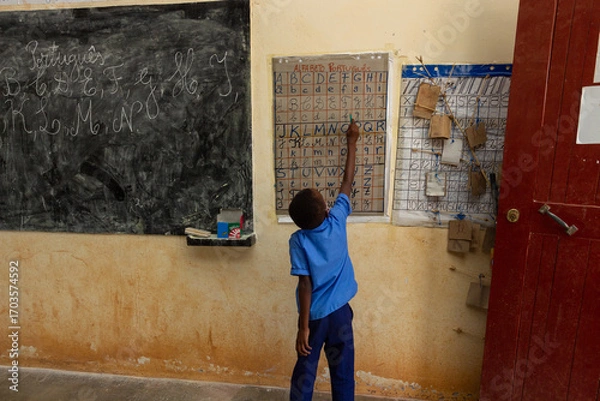 Fototapeta A young African boy in a blue shirt points to an alphabet chart on a wall in a simple, sparse classroom, learning to read and write.