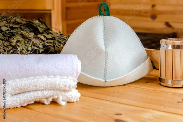 Fototapeta Sauna equipment. Bucket with ladle, felt wool hat, birch whisk and towels on wooden table indoors
