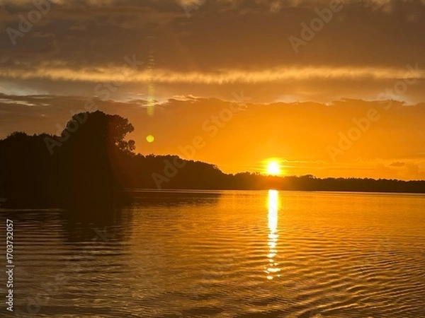 Obraz Summer sunset landscape sun setting over Norfolk Broads England uk tranquil water with the beautiful orange yellow blue skies viewed from sailing boat to horizon and trees reeds, clouds reflected 