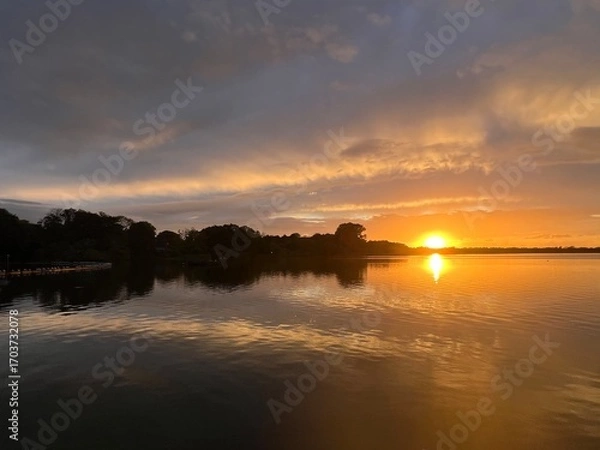 Fototapeta Summer sunset landscape sun setting over Norfolk Broads England uk tranquil water with the beautiful orange yellow blue skies viewed from sailing boat to horizon and trees reeds, clouds reflected 