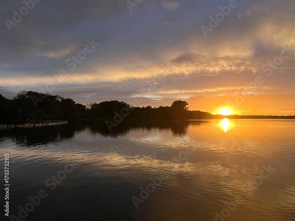 Fototapeta Summer sunset landscape sun setting over Norfolk Broads England uk tranquil water with the beautiful orange yellow blue skies viewed from sailing boat to horizon and trees reeds, clouds reflected 