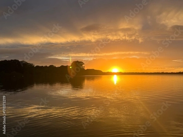 Obraz Summer sunset landscape sun setting over Norfolk Broads England uk tranquil water with the beautiful orange yellow blue skies viewed from sailing boat to horizon and trees reeds, clouds reflected 