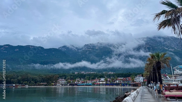Fototapeta Scenic view of Akyaka, Turkey, with misty mountains, palm trees, and waterfront promenade. Atmospheric cloudy landscape with seaside town, nature, and coastal beauty.