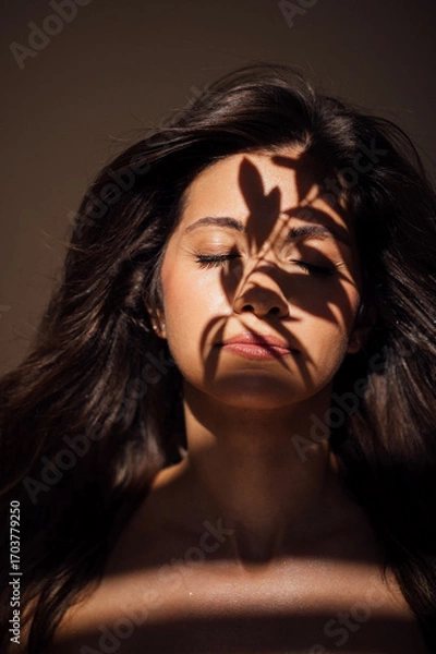 Fototapeta Young woman with long dark hair, eyes closed, illuminated by soft light, with intricate shadows cast on her face, creating a serene and contemplative atmosphere in a minimalist setting