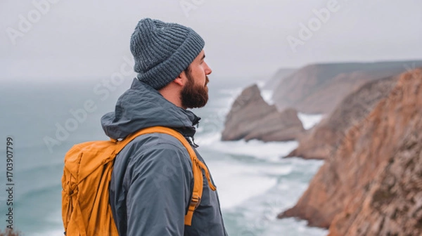 Fototapeta Man wearing a beanie gazes at rugged coastal cliffs while standing on a rocky outcrop by the ocean during an overcast day
