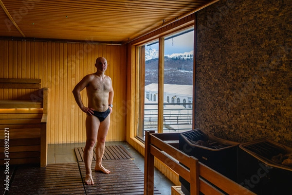 Fototapeta A sporty man in swimming trunks stands by the window in a sauna with a gorgeous view of the mountains.