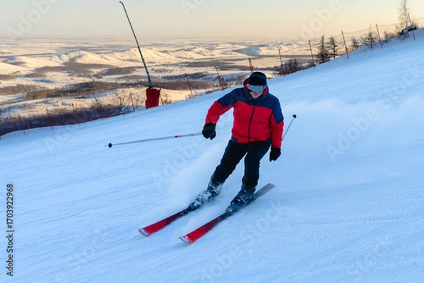 Fototapeta A man skis at high speed in a red ski suit, glasses, with poles and skis. Slowing down on turns, snowdrifts form behind him.