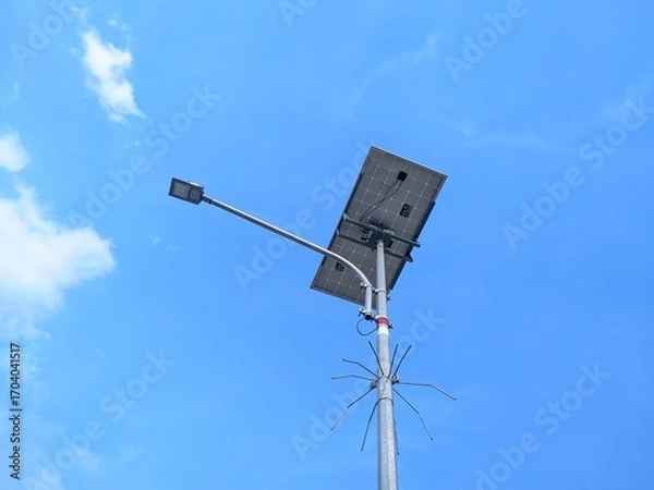 Fototapeta Solar-powered street light with a solar panel and LED lamp against a bright blue sky with scattered clouds.