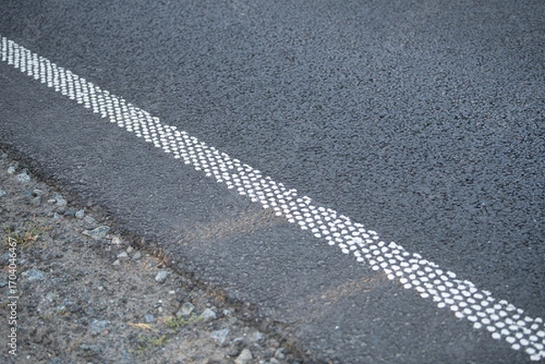 Fototapeta A close view of a black asphalt road with textured white dotted edge markings. The natural terrain is visible alongside the road, showing gravel and grass patches