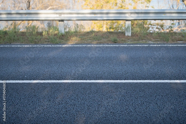 Fototapeta A clear view of a well-maintained asphalt road with a white striped line and a guardrail. The background features trees with leaves, indicating a sunny day