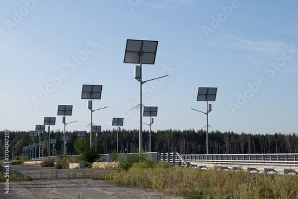 Fototapeta Rows of solar panels stand along an abandoned road, showcasing renewable energy technology in a quiet, forested location under the bright sky. The area appears untouched and serene