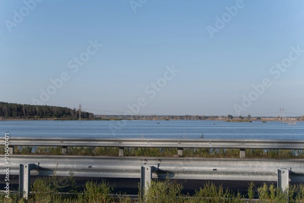 Fototapeta A serene view showcasing calm waters beside a highway, with lush greenery lining the roadside and trees in the distance under a clear blue sky