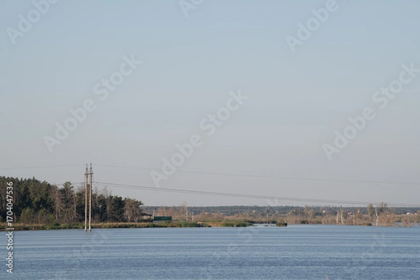 Fototapeta A serene river flows gently with a clear horizon. Soft light casts shadows on the distant trees and utility poles, creating a peaceful atmosphere near the water's edge