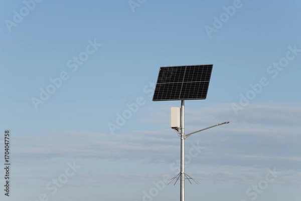 Fototapeta A solar panel is mounted on a tall metal pole, positioned in an open area under a bright blue sky. The environment is calm and sunny, highlighting renewable energy equipment