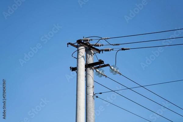 Fototapeta A utility pole stands tall with power lines stretching across a bright blue sky. The scene captures the infrastructure essential for electricity distribution