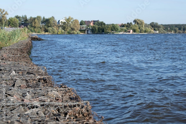 Fototapeta A peaceful riverbank extends along the water, featuring gentle waves and vibrant greenery. A small town is visible in the background under clear blue skies