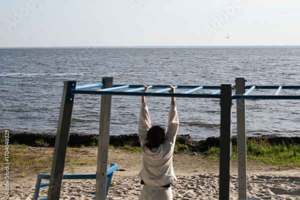 Fototapeta A woman hangs from a horizontal bar on a beach workout station, enjoying the peaceful ocean view on a clear day. Waves gently lap at the shore, enhancing the serene atmosphere
