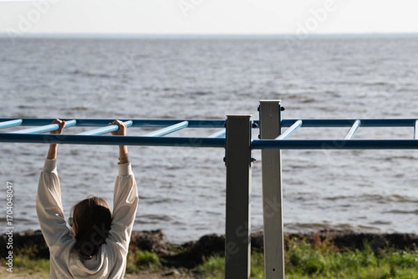 Fototapeta A person hangs from a blue exercise bar overlooking the calm ocean water. The scene captures a serene moment of fitness and nature under clear skies