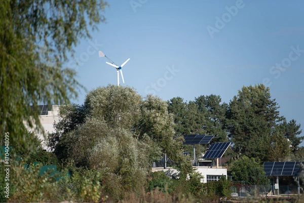 Fototapeta A wind turbine spins atop a green hill while nearby solar panels glisten in the sun on rooftops. Trees provide shade in this eco-friendly residential neighborhood under a blue sky