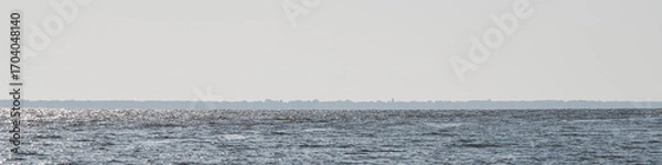 Fototapeta A serene ocean stretches towards a distant shoreline under a bright sky. The water sparkles in the sunlight, creating a peaceful atmosphere typical of a midday scene
