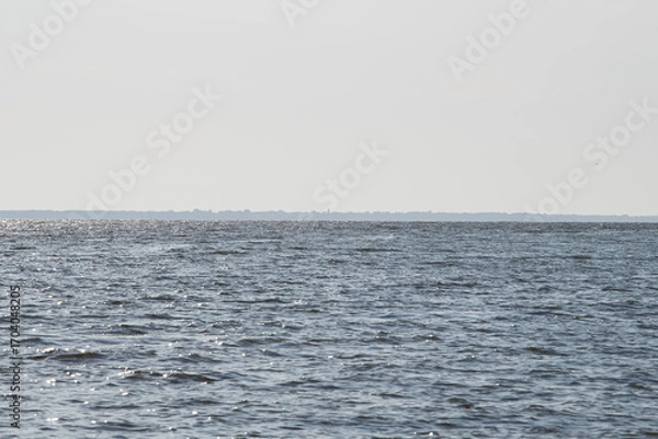 Fototapeta Gentle waves ripple across the water as the sun reflects off the surface. In the background, the outline of a distant shoreline is visible, creating a serene atmosphere