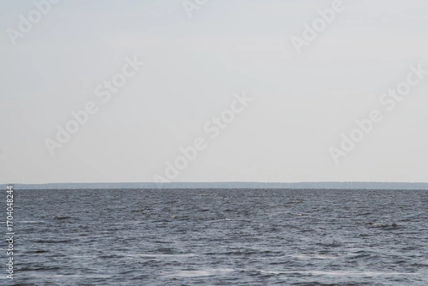 Fototapeta The tranquil water spreads out towards the horizon, reflecting a clear sky. A distant shoreline is subtly visible, creating a serene atmosphere