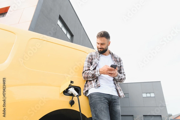 Fototapeta Delivery driver using smartphone while charging electric van