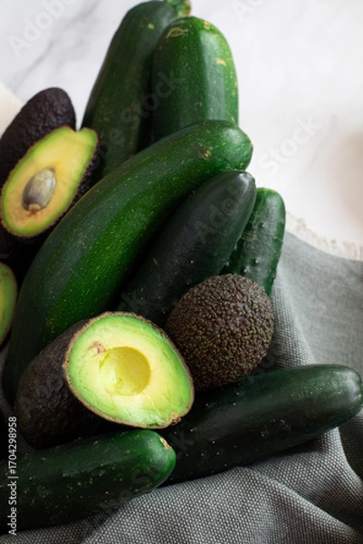 Obraz Avocados, cucumbers, and zucchinis arranged on gray fabric with a white marble background.