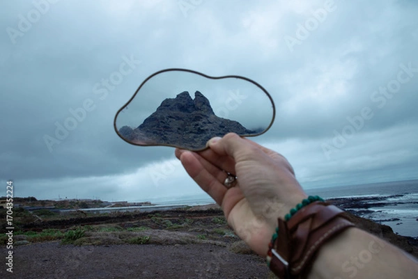 Obraz A hand holds a cloud-shaped mirror reflecting a mountain peak, with a cloudy ocean backdrop 