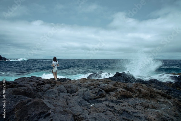 Obraz Woman standing on volcanic rocks near crashing ocean waves. Overcast sky and wild seascape create a dramatic coastal scene.