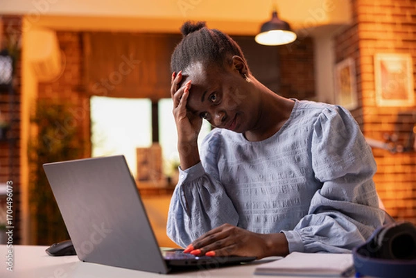 Fototapeta Overworked businesswoman in apartment, showing evening fatigue during remote work. Stressed and upset freelancer rubs forehead with one hand while typing on laptop, struggling to manage schedule.
