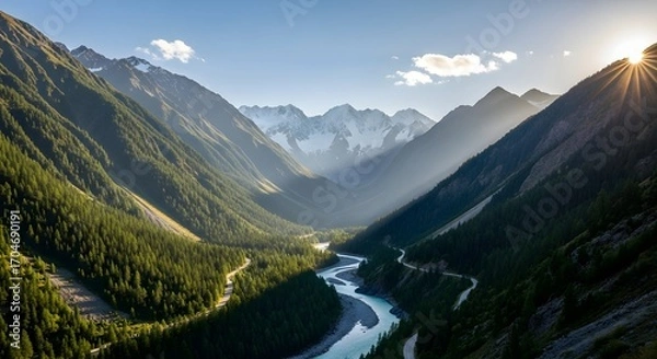 Fototapeta Sunburst over a winding river valley with dense pine forests and snow capped mountains