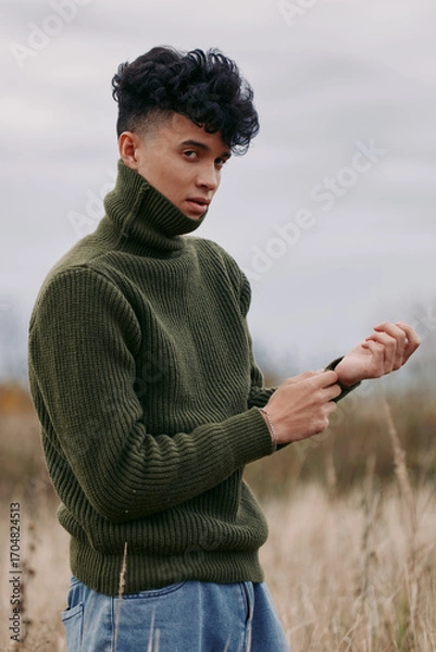 Fototapeta A young man in a textured olive turtleneck stands in a windswept field, posing naturally with a calm gaze and genuine posture that communicates authentic, credible everyday style.