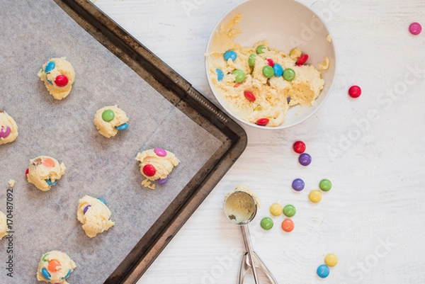 Fototapeta Cookie dough with colorful chocolate candies in bowl next to dough scooper and scoops of cookie dough on baking sheet