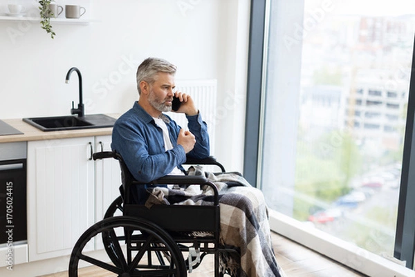 Fototapeta Senior Caucasian man sitting in wheelchair by kitchen indoors using phone near window. Scene depicts everyday lifestyle, accessibility, comfort, connection, communication, independence, and inclusion