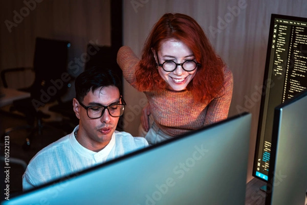 Fototapeta Two coworkers collaborate at dual monitors in a dim office, reviewing code and brainstorming solutions during late night software development.