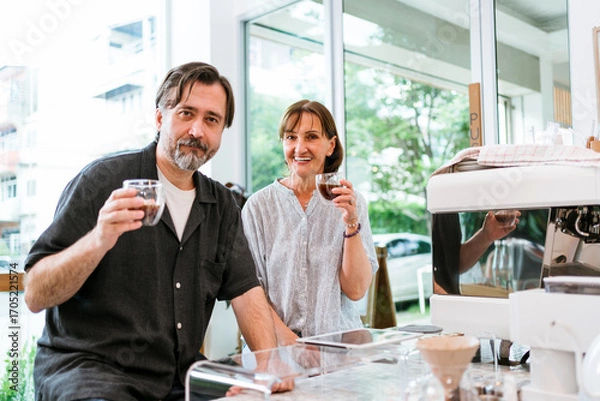 Fototapeta Smiling adults enjoying espresso in a bright modern cafe beside a professional coffee machine, raising cups at the counter with natural light and urban view through large windows.