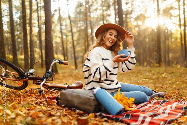 Obraz A happy woman sits on a red blanket next to a bicycle, drinking hot tea from a thermos. A young woman relaxes in a clearing in an autumn park. Picnic and cycling concepts