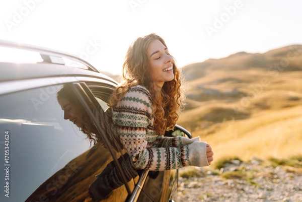 Obraz A happy traveler leans out of a car window at sunset. The young woman feels free and enjoys the mountain scenery during her drive. Adventure and transportation concept. Active lifestyle.